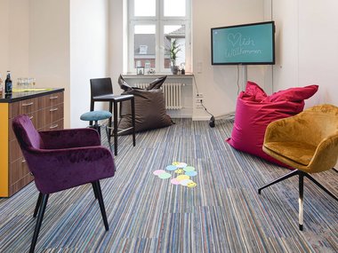 Ein schmaler Tagungsraum mit verschiedenen, bunten Stühlen und einem Bildschirm