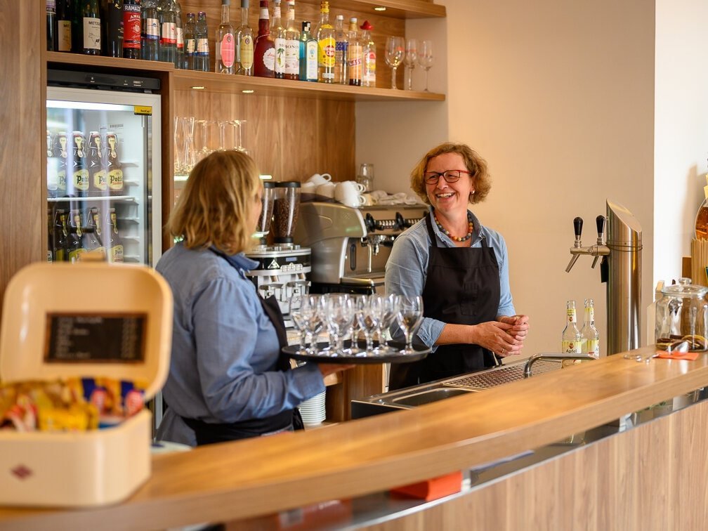 Zwei Servicemitarbeiterinnen hinter der Theke, eine trägt ein Tablett mit Wassergläsern 