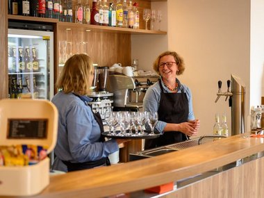 Zwei Servicemitarbeiterinnen hinter der Theke, eine trägt ein Tablett mit Wassergläsern 